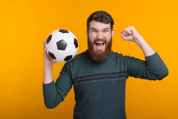 Uomo barbuto emozionante che tiene una palla in mano mentre screamin e facendo gesto del vincitore sullo spazio giallo.
