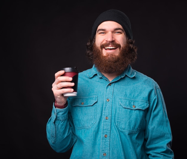 Uomo barbuto felice che tiene tazza di caffè per andare su sfondo scuro