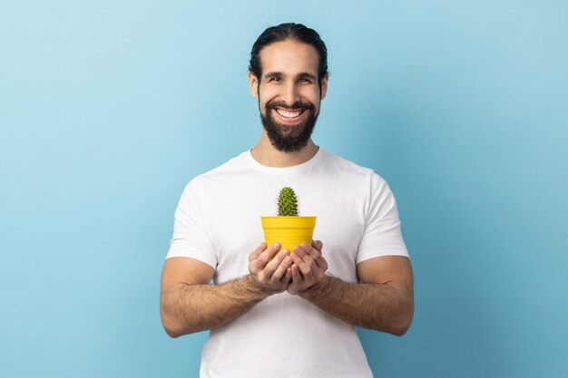 Uomo barbuto felice che tiene vaso di fiori gialli e cactus guardando la fotocamera con un sorriso a trentadue denti