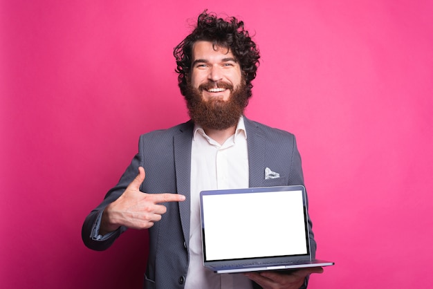 Uomo barbuto felice in vestito che indica allo schermo bianco vuoto sul computer portatile e sorridente