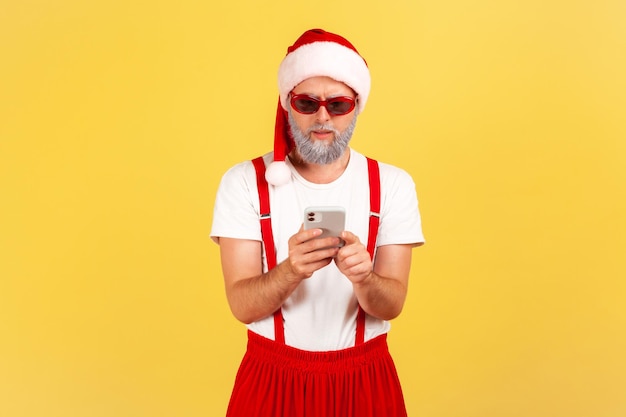 Uomo barbuto grigio concentrato serio in occhiali da sole e cappello rosso di Babbo Natale che tiene smartphone e digita un messaggio o legge e-mail Colpo in studio al coperto isolato su sfondo giallo