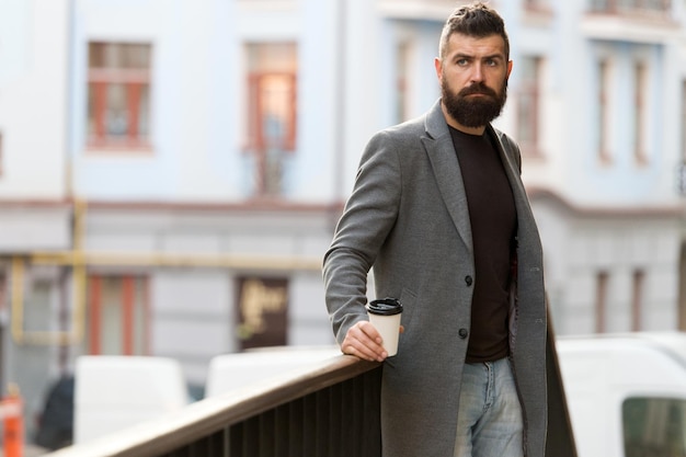 Uomo barbuto hipster bere caffè tazza di carta. Un altro sorso di caffè. Stile di vita della città. L'aspetto ben curato dell'uomo d'affari gode della pausa caffè dal fondo urbano del centro affari. Rilassati e ricaricati.