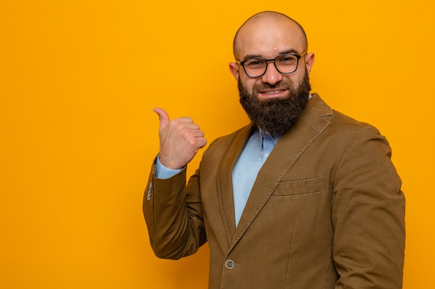 Uomo barbuto in abito marrone con gli occhiali che sembra felice e positivo sorridente allegramente rivolto indietro con il pollice