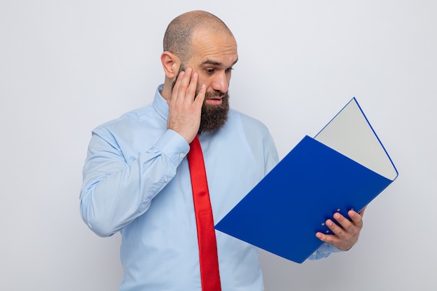 Uomo barbuto in cravatta rossa e camicia blu che tiene la cartella dell'ufficio guardandolo confuso e sorpreso in piedi su sfondo bianco