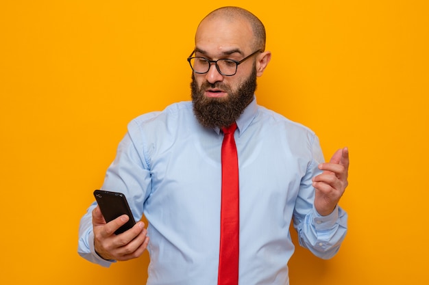 Uomo barbuto in cravatta rossa e camicia blu con gli occhiali che tengono lo smartphone guardandolo sorpreso e confuso