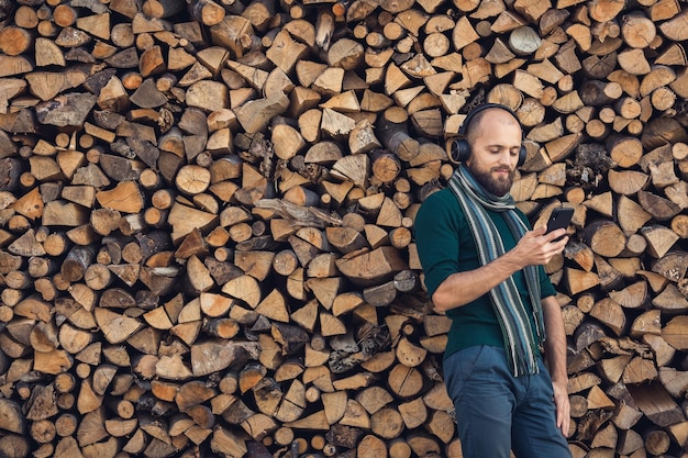 Uomo barbuto in cuffie bluetooth che ascolta musica all'aperto vicino a un mucchio di legna da ardere