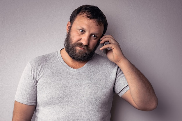Uomo barbuto in maglietta grigia che tiene il telefono cellulare mentre in piedi contro il muro grigio