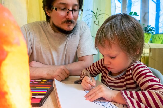 Uomo barbuto in maglietta grigia e ragazzo carino che disegnano immagini con pastelli a cera colorati mentre trascorrono del tempo a casa insieme