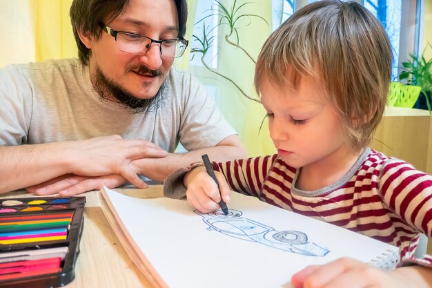 Uomo barbuto in maglietta grigia e ragazzo carino che disegnano immagini con pastelli a cera colorati mentre trascorrono del tempo a casa insieme