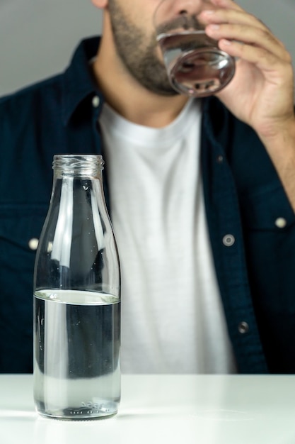 Uomo barbuto irriconoscibile che beve acqua da un bicchiere di vetro con una bottiglia di vetro mezza piena in primo piano concetto di salute e idratazione