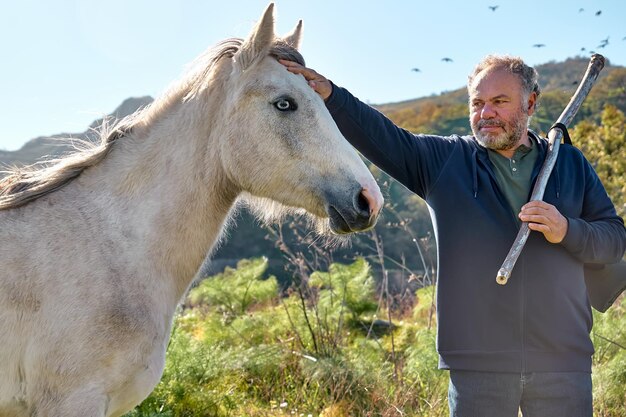 Uomo barbuto maturo che incontra il cavallo bianco mentre fa un'escursione nel pascolo rurale Concetto di amicizia e relazione Benessere e unità con la natura Strada per la montagna