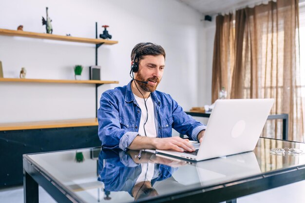 Uomo barbuto maturo in cuffie che lavorano al tavolo con il computer portatile in ufficio