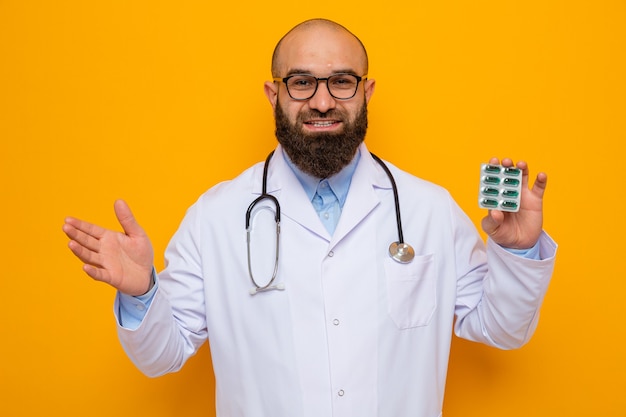 Uomo barbuto medico in camice bianco con stetoscopio intorno al collo con gli occhiali che tengono blister con pillole guardando la fotocamera felice e positivo sorridente in piedi su sfondo arancione