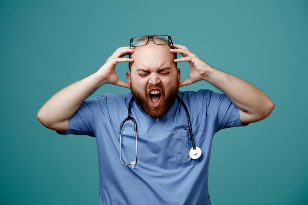 Uomo barbuto medico in uniforme con uno stetoscopio intorno al collo con gli occhiali arrabbiato e frustrato che urla tirandosi i capelli impazziti in piedi su sfondo blu