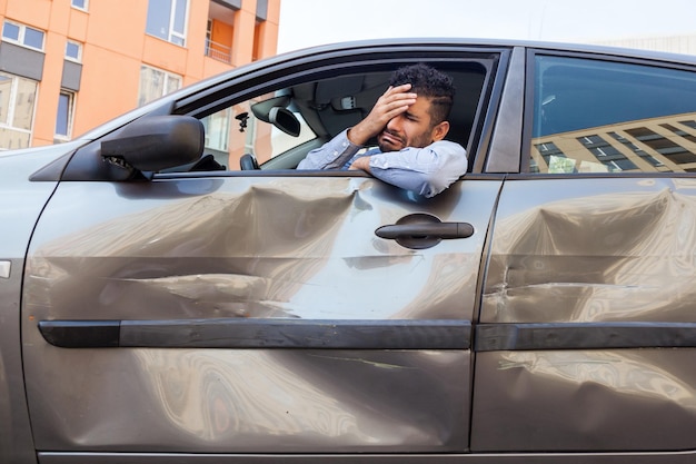 Uomo barbuto piangente che indossa una camicia blu seduto al volante in un'automobile distrutta e copre metà del viso con le palme disperazione dopo che l'incidente stradale ha bisogno di riparare la porta automatica Scatto all'aperto