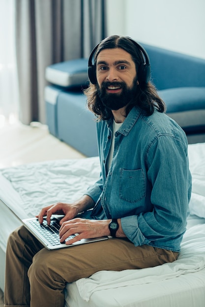 Uomo barbuto positivo che indossa le cuffie a casa e sorride mentre è seduto con un laptop moderno