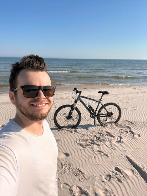 Uomo barbuto positivo che indossa occhiali da sole sorridenti davanti alla sua bicicletta sulla spiaggia Concetto di sport e viaggio