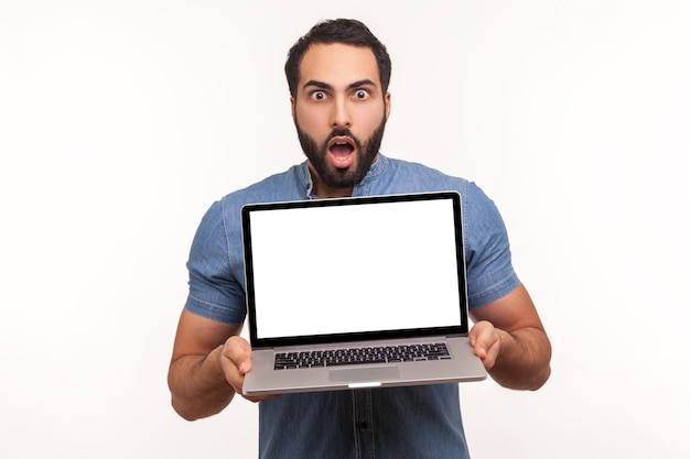 Uomo barbuto scioccato che mostra laptop con display vuoto sorpreso con area pubblicitaria guardando la fotocamera con occhi grandi e bocca aperta Colpo in studio indoor isolato su sfondo bianco