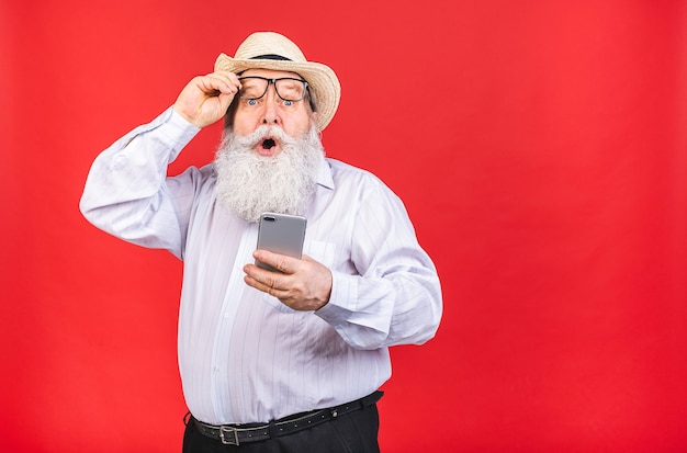 uomo barbuto scioccato con cappello e occhiali in possesso di un telefono cellulare isolato su sfondo rosso