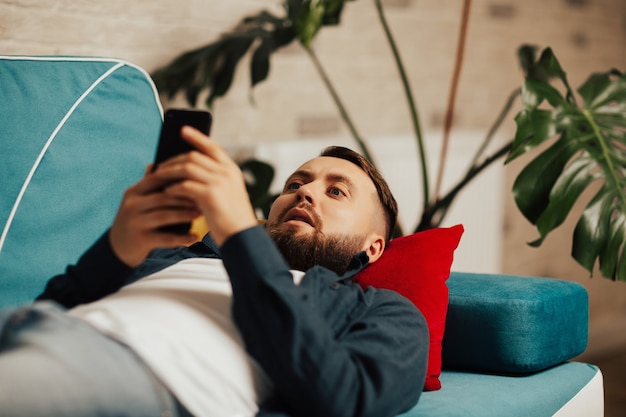 uomo barbuto sdraiato sul divano di casa utilizzando internet sul suo telefono cellulare e guardando sorpreso e scioccato.