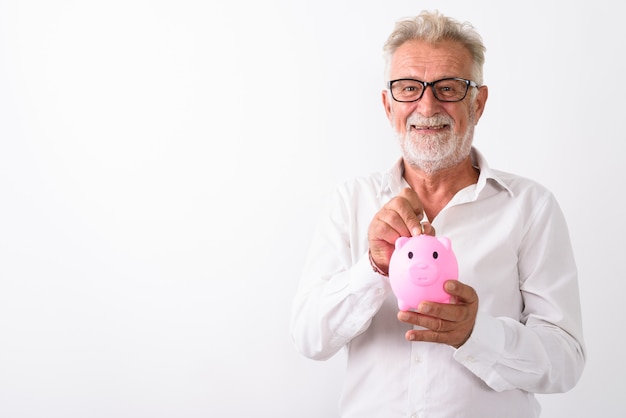 uomo barbuto senior felice che sorride mentre mette moneta nel salvadanaio con gli occhiali su bianco