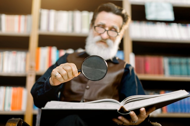 Uomo barbuto sorridente bello, bibliotecario o professore, in biblioteca, seduto sullo sfondo di librerie, con lente d'ingrandimento e libro di lettura. Concentrati sul vetro e sul libro