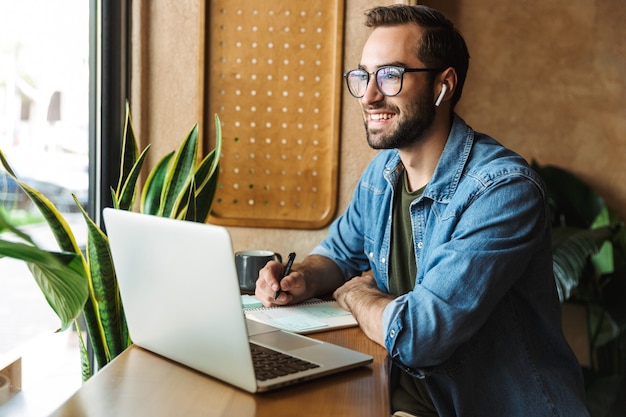 uomo barbuto sorridente che indossa occhiali scrivendo note e usando il laptop mentre si lavora in un bar al chiuso