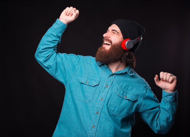 Uomo barbuto sorridente felice che ascolta musica alle cuffie senza fili e balla sopra il muro scuro