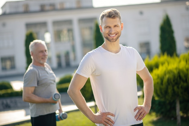 Uomo barbuto sorridente, mani sui fianchi, uomo dai capelli grigi con manubri dietro