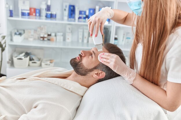 Uomo barbuto sorridente nel salone di bellezza professionale spa durante la procedura di pulizia del viso ad ultrasuoni.