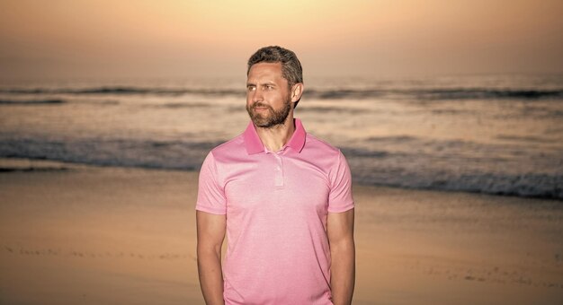 Uomo barbuto sorridente sopra il mare sulla moda estiva della spiaggia di tramonto d'estate