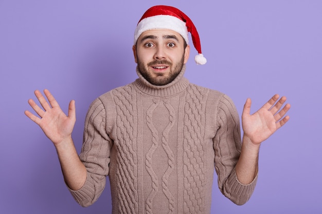 Uomo barbuto stupito che indossa cappello da Babbo Natale e pullover beige in piedi con la bocca aperta e grandi occhi