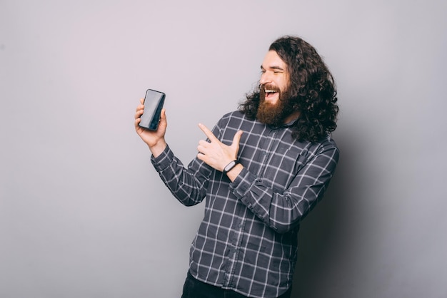 Uomo barbuto stupito con i capelli lunghi che punta allo schermo dello smartphone in piedi su uno sfondo grigio