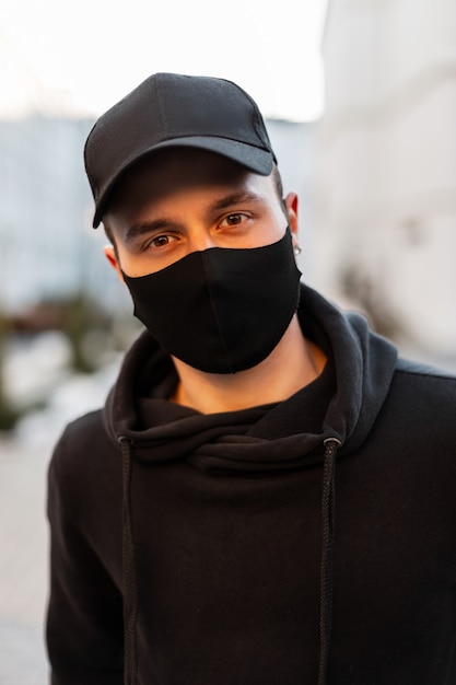 Uomo bello alla moda con berretto nero e maschera medica con felpa con cappuccio alla moda per strada. concetto di stile casual maschile pandemico e urbano