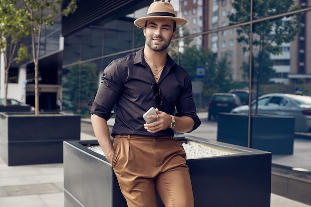 Uomo bello brutale abbronzato muscoloso hipster in camicia e cappello neri con il telefono