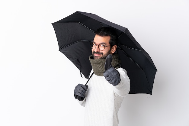 Uomo bello caucasico con la barba che tiene un ombrello sopra la parete bianca con i pollici su perché è successo qualcosa di buono