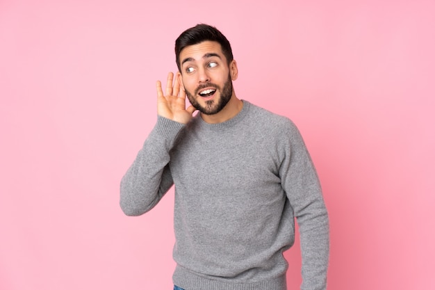 Uomo bello caucasico sopra la parete isolata che ascolta qualcosa mettendo la mano sull'orecchio