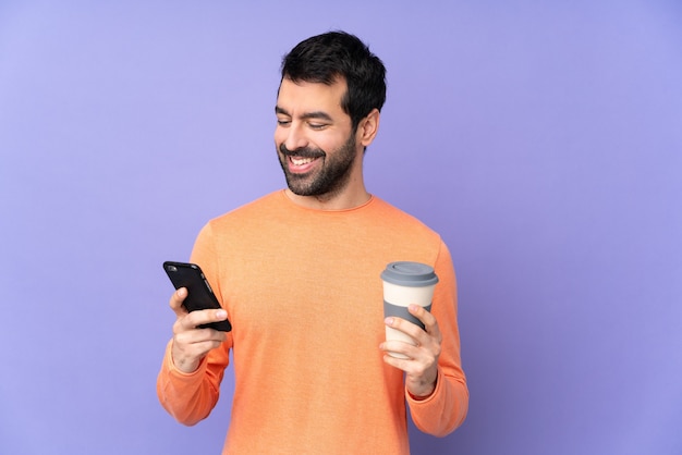 Uomo bello caucasico sopra la parete viola isolata che tiene caffè da portare via e un cellulare