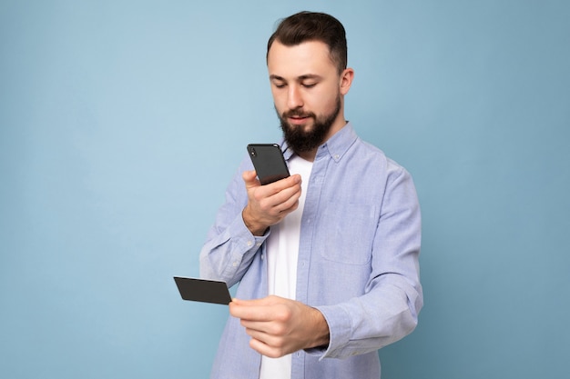 Uomo bello che indossa abiti di tutti i giorni isolati sulla parete di fondo tenendo e utilizzando il telefono e la carta di credito per effettuare il pagamento guardando lo schermo dello smartphone,