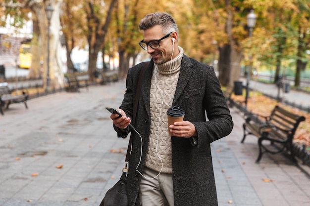 Uomo bello che indossa un cappotto che cammina all'aperto, ascoltando musica con auricolari e telefono cellulare, tenendo la tazza di caffè da asporto