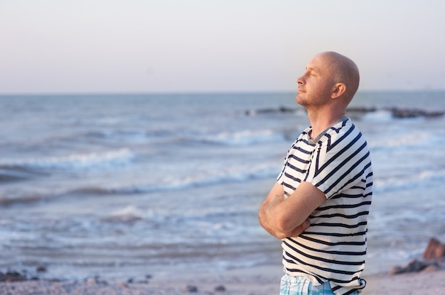 Uomo bello che indossa una maglietta a righe che respira davanti all'oceano sulla spiaggia.