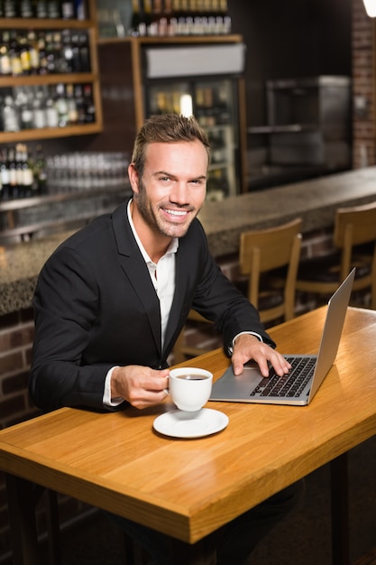 Uomo bello che per mezzo del computer portatile e mangiando un caffè