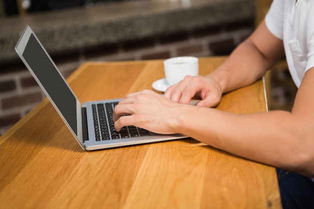 Uomo bello che per mezzo del computer portatile e mangiando un caffè