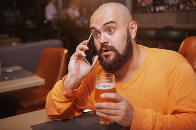 Uomo bello che sembra sorpreso, parlando al telefono bevendo birra al ristorante