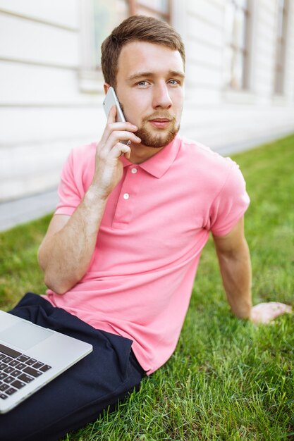 Uomo bello che si siede sull'erba nella città con un computer portatile e che parla sul telefono, ricerca di lavoro