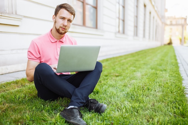 Uomo bello che si siede sull'erba nella città con un computer portatile, ricerca di lavoro