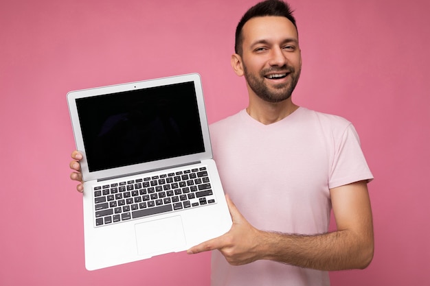 Uomo bello che tiene il computer portatile che guarda l'obbiettivo in t-shirt su sfondo rosa isolato