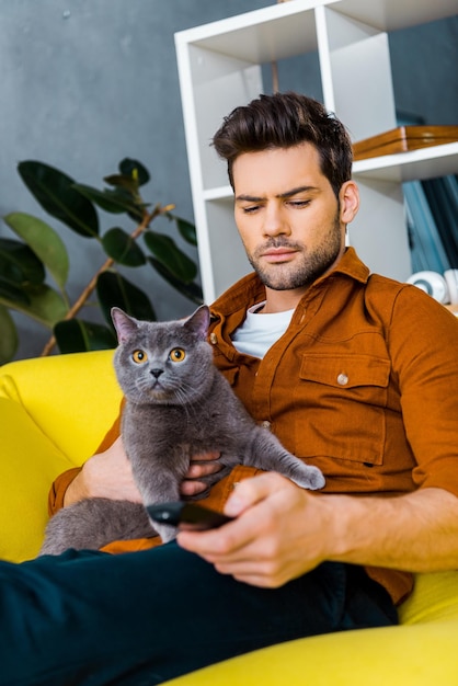 Uomo bello che tiene telecomando e gatto grigio a casa