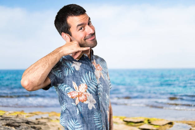 Uomo bello con camicia di fiori che fanno gesto di telefono in spiaggia