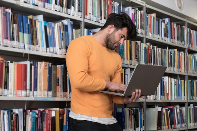 Uomo bello con capelli scuri in piedi nella biblioteca Laptop e organizer sul tavolo guardando lo schermo un concetto di studiare libri sfocati sul retro